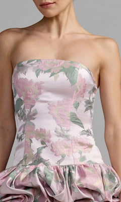 A woman wearing the Mac Duggal 69156 Short Party Dress, which features a strapless fitted bodice and a ruffled skirt. Made from floral brocade fabric with pink flowers and green leaves on a light background, this dress offers a fresh, feminine look.