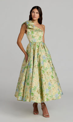 A woman stands with one hand in her pocket against a plain background, wearing the Tea-Length Party Dress: Mac Duggal 69160 in Green Multi—a striking one-shoulder style adorned with vibrant floral print and finished with a large bow on the shoulder.