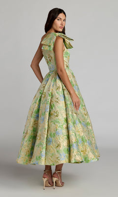 A woman wears the Mac Duggal 69160 Tea-Length Party Dress, featuring a floral pattern, full skirt, and bow detail at the shoulder, styled with neutral heels against a plain background.