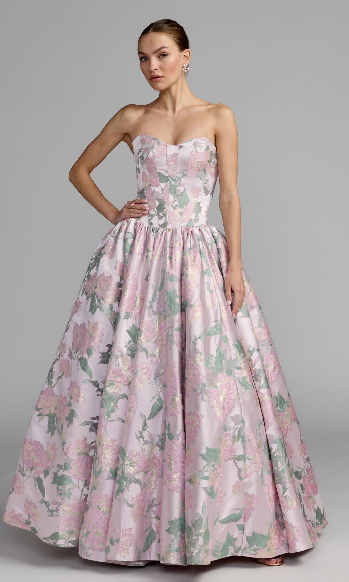 A woman poses against a plain gray background wearing the Mac Duggal 69162 long formal dress in Pink Multi, a strapless ball gown with a full skirt and an eye-catching metallic floral print in pink and green shades.
