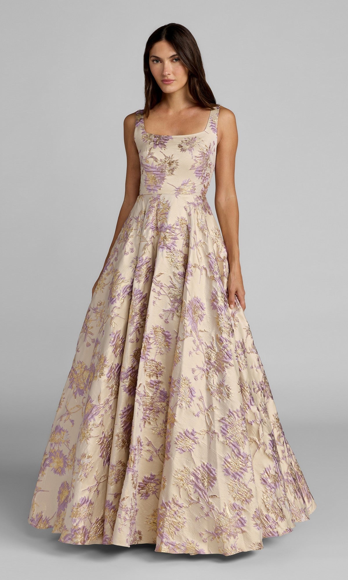 A woman wearing the Mac Duggal 69163 long formal dress in Champagne Multi, a sleeveless, floor-length gown featuring a floral brocade design, stands against a plain gray background.