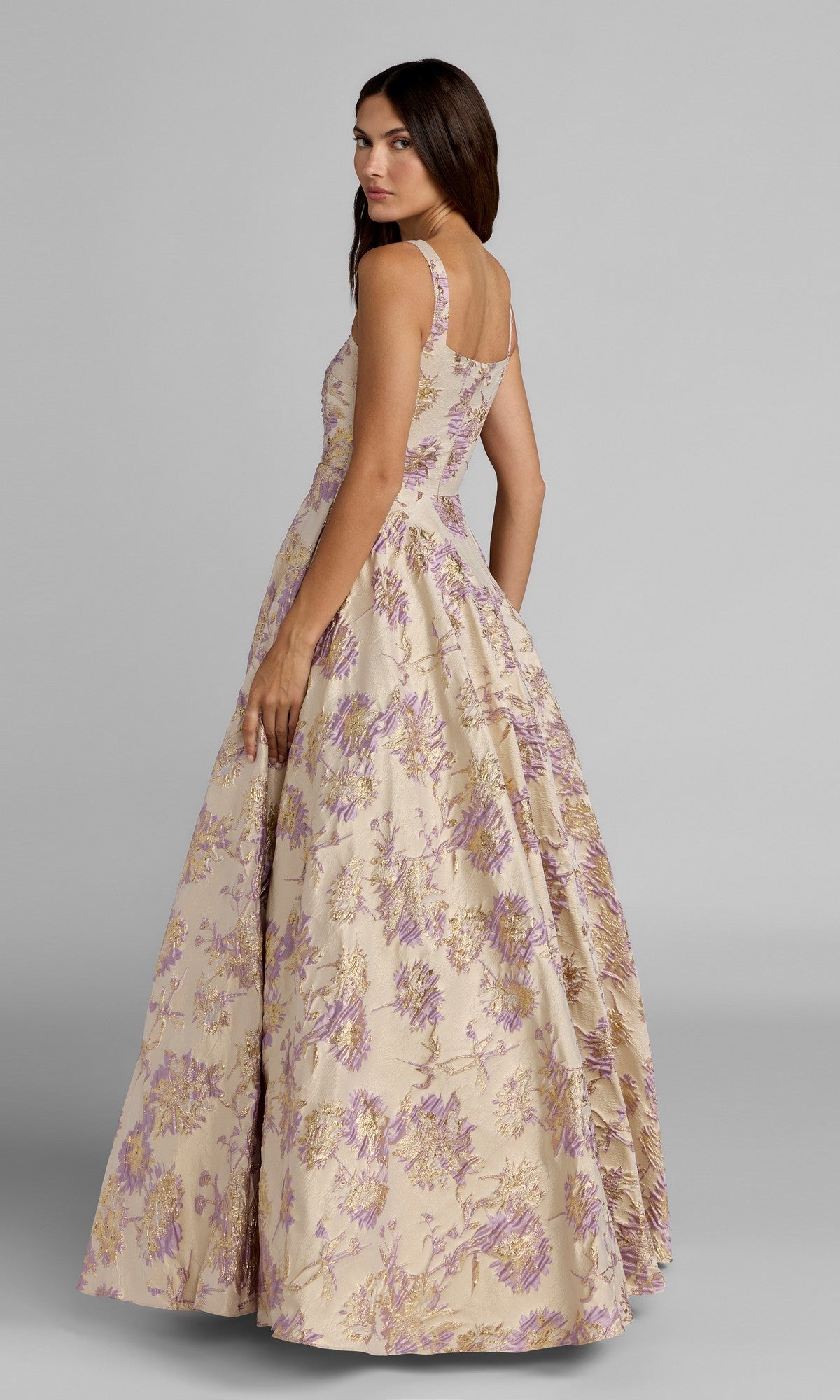 A woman wearing the Long Formal Dress: Mac Duggal 69163, featuring a sleeveless design, full ballgown skirt, and delicate floral-print pattern, looks over her shoulder against a plain gray background.