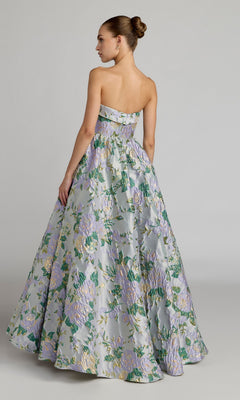 A woman stands with her back to the camera against a plain backdrop, wearing the Mac Duggal 69172—a strapless, pastel floral ballgown with metallic accents—her hair styled in a sleek bun.