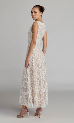 A woman stands in profile against a plain gray background, wearing the Mac Duggal 69192 tea-length party dress—an elegant sleeveless lace design with an A-line skirt—paired with silver high-heeled sandals.