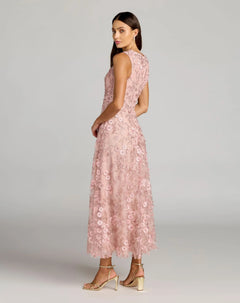 A woman stands sideways against a plain gray background, wearing the Mac Duggal 69192 tea-length party dress—a sleeveless light pink ensemble with a floral lace overlay, fitted bodice, and elegant A-line skirt—paired with ankle-strap heels.