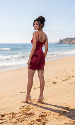 A woman wearing the Atria 6926S short homecoming dress in red stands on a sandy beach near the shoreline, facing away from the camera with cliffs visible in the distance.