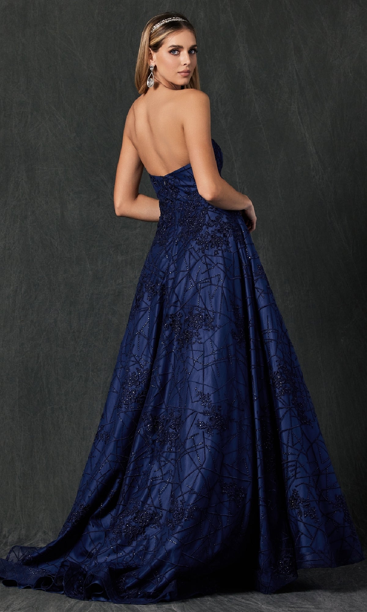 A woman poses with her back slightly turned against a dark textured backdrop, wearing the Juliet 692—a floor-length, strapless navy blue prom dress featuring embroidered patterns and a subtle sweetheart neckline.