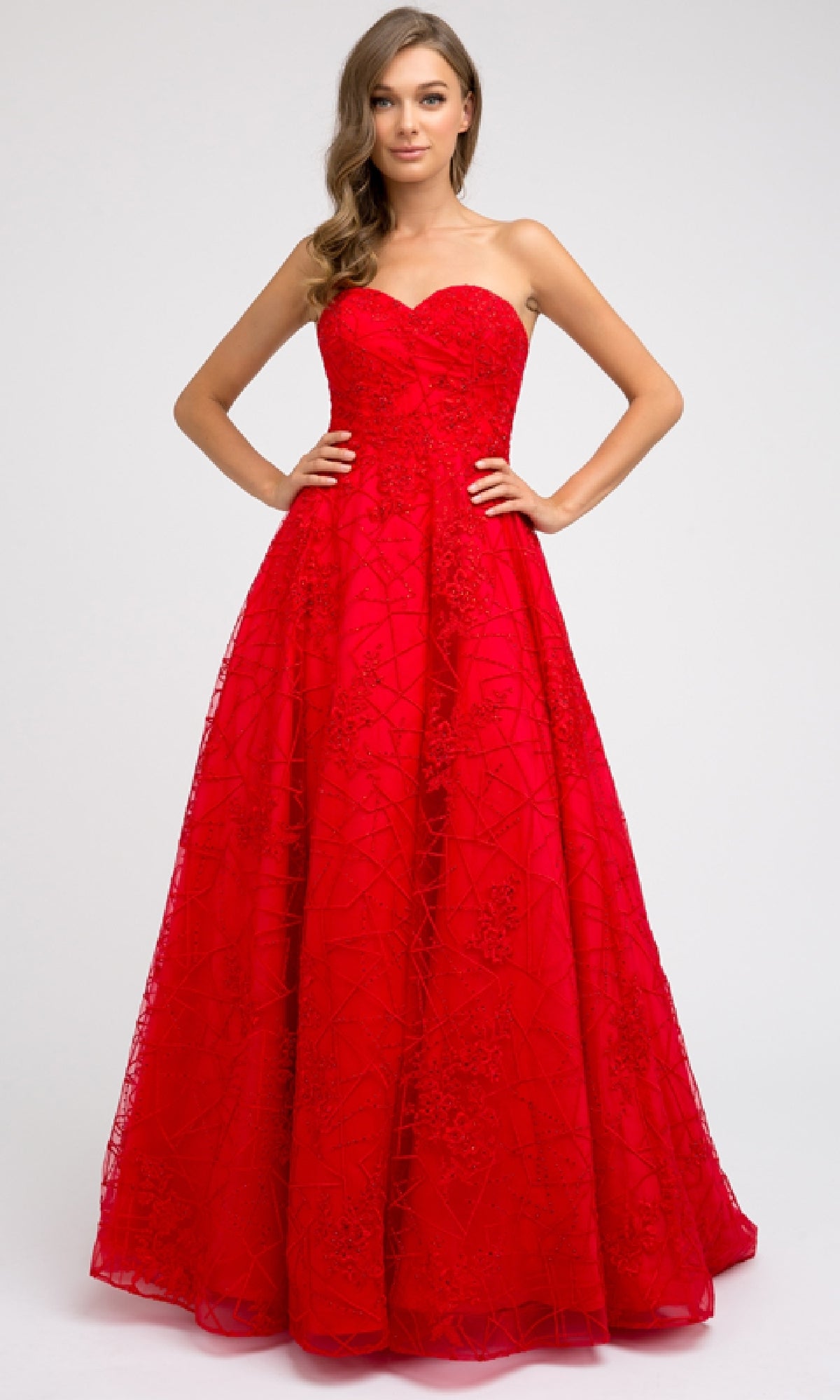 A woman wears the Juliet 692 long prom dress, a strapless red gown with a sweetheart neckline, fitted bodice, lace embellishments, and floor-length skirt, posing with hands on hips against a plain light background.