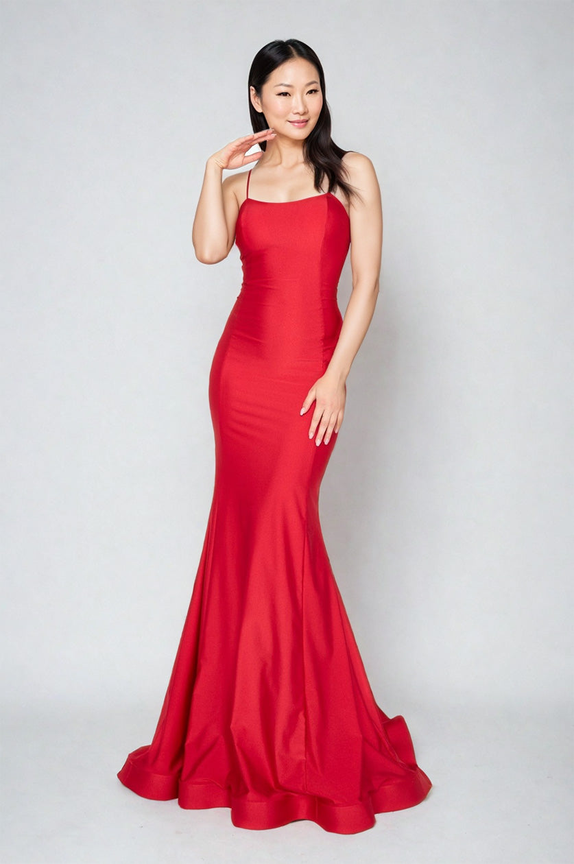 A woman poses against a plain backdrop in the Atria 6520H Lace-Up Back Long Prom Dress, a sleeveless, floor-length red gown. Her hair is down, and she raises one hand near her face.