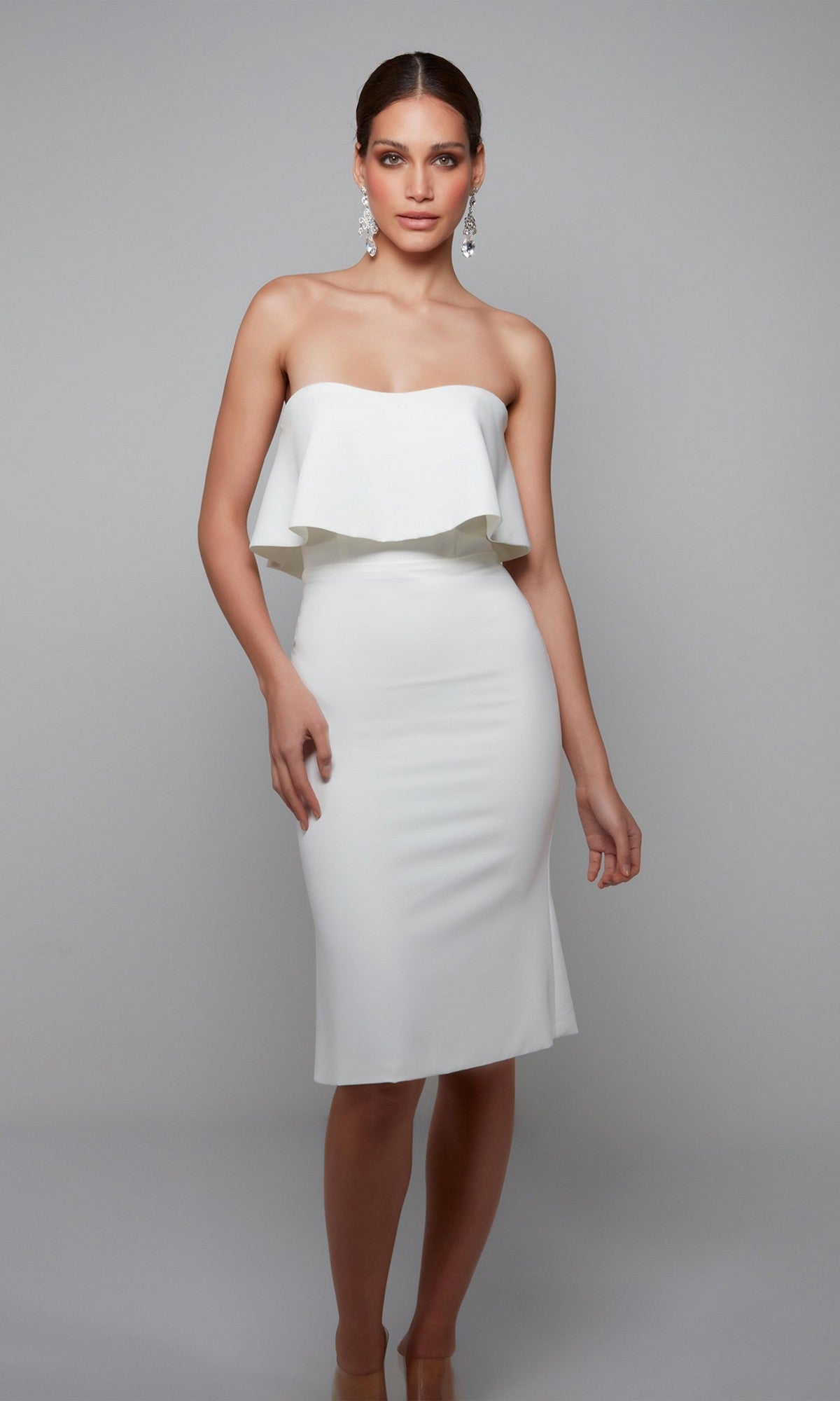A woman in an ivory Alyce 70002 knee length semi-formal dress with a ruffled top stands against a plain gray background.