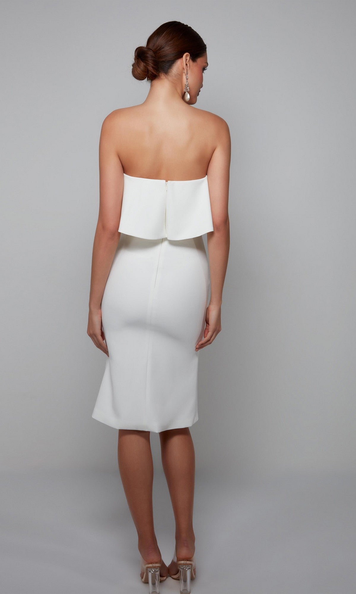 A woman stands against a plain background wearing the Knee Length Semi-Formal Dress: Alyce 70002 in white, paired with clear high heels; her hair is styled in a low bun.