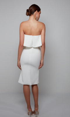 A woman stands against a plain background wearing the Knee Length Semi-Formal Dress: Alyce 70002 in white, paired with clear high heels; her hair is styled in a low bun.
