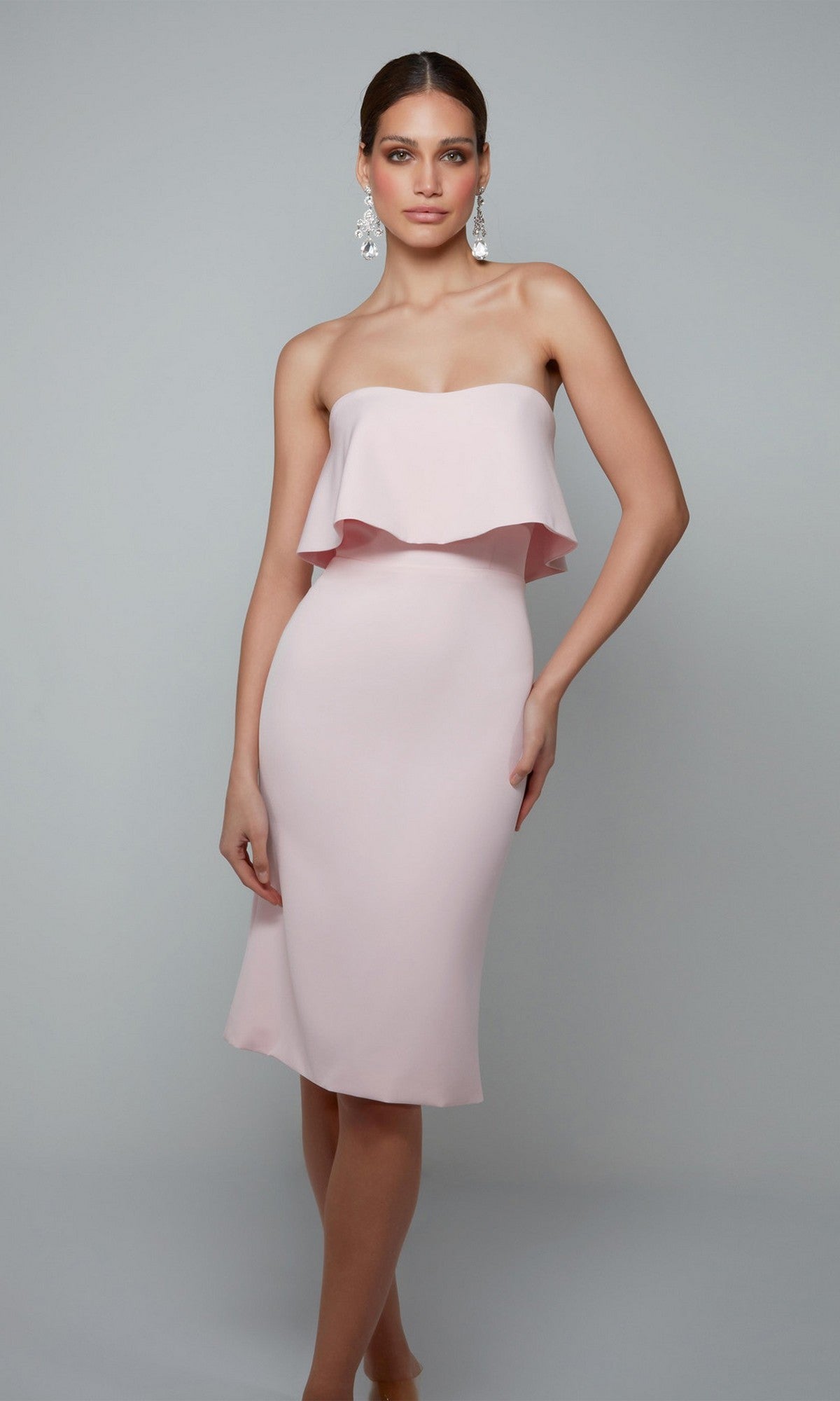 A woman modeling the Knee Length Semi-Formal Dress: Alyce 70002 in Rosewater, featuring a strapless design and ruffled overlay, stands against a plain gray background.