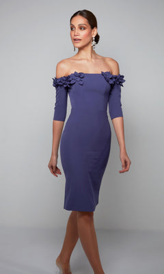 A woman stands against a plain gray background wearing the Alyce 70003, a fitted, off-the-shoulder purple knee-length semi-formal dress with floral embellishments on the neckline.