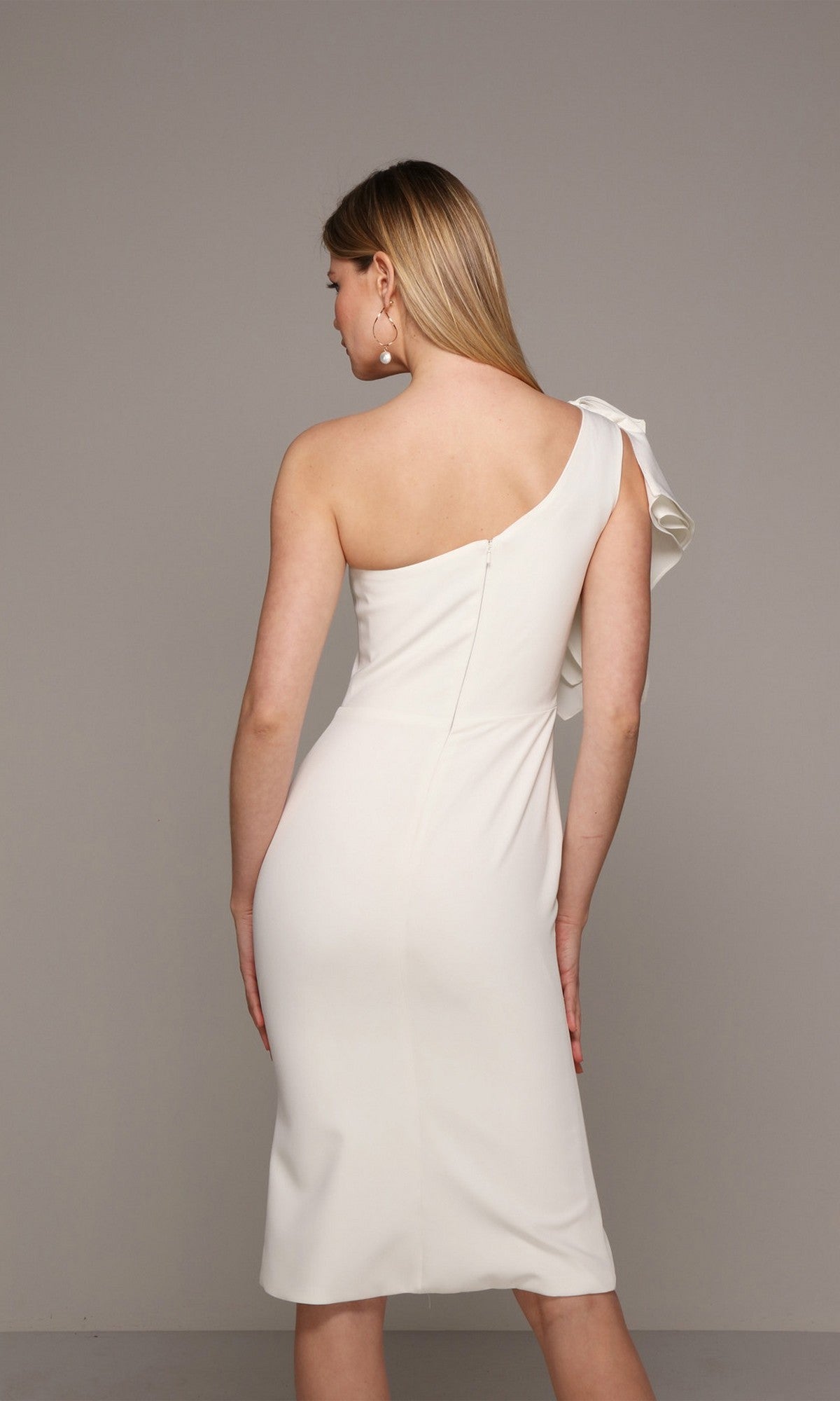 A woman stands facing away against a plain background, modeling the Alyce 70005 knee-length semi-formal dress—a white, one-shoulder design with ruffle detail.