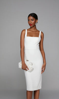 Woman wearing the knee length semi-formal Alyce 70010 dress in ivory, holding a white floral clutch, standing against a plain gray background.