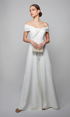 A woman wearing the Long Formal Jumpsuit: Alyce 70013 in off-the-shoulder white holds a textured beige clutch while standing against a plain light gray background.