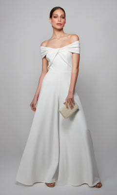 A woman stands against a plain background wearing the Alyce 70013 long formal jumpsuit, paired with a beaded clutch and sandals.