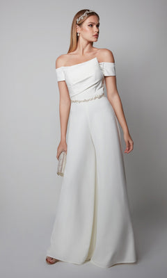 Woman wearing the Long Formal Jumpsuit: Alyce 70014, featuring an off-the-shoulder design and wide legs, paired with a small clutch and jeweled headband and earrings against a plain background.