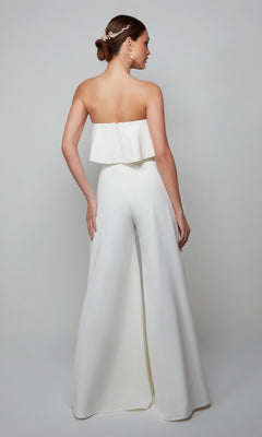 A woman poses with her back to the camera against a plain background, wearing the Long Formal Jumpsuit: Alyce 70015. The strapless jumpsuit features wide-leg pants, a layered top, and she styles her low bun with a decorative hair accessory.