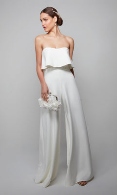 A woman in the Long Formal Jumpsuit: Alyce 70015, featuring wide-leg pants, holds a white floral clutch and has her hair styled in an updo adorned with a hair accessory.