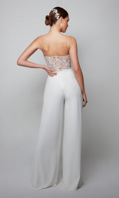 Woman facing away in the Long White Jumpsuit: Alyce 70017, which has a white lace bodice, backless design with spaghetti straps, and wide-leg pants. Her hair is styled in a bun with a decorative accessory.