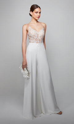 A woman wears the Long White Jumpsuit: Alyce 70017, which features a white lace bodice and wide-leg pants, holding a white floral clutch against a plain light background.