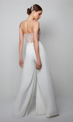 A woman poses gracefully with her back turned, wearing the Alyce 70017 long white jumpsuit featuring a sheer lace bodice and wide-leg pants.