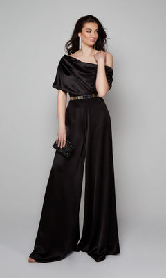 A woman models the Alyce 70019 long formal jumpsuit in black, featuring an off-shoulder neckline and wide-leg pants, paired with a metallic belt, long earrings, and a black clutch against a plain background.