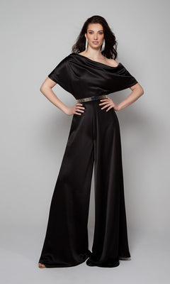 A woman poses in a studio against a plain background wearing the Long Formal Jumpsuit: Alyce 70019, a black wide-leg jumpsuit with short sleeves and a metallic belt, hands on hips.