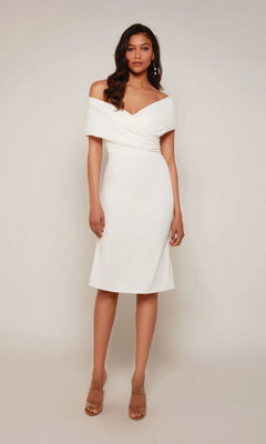 A woman faces forward in the Alyce 70023 knee-length white party dress with a classic sheath silhouette and beige heeled sandals, standing against a plain light background.