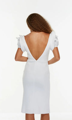 A woman with curly hair models the Alyce 70024 knee length white party dress, which has ruffled short sleeves and a deep V-back, standing against a plain white background.