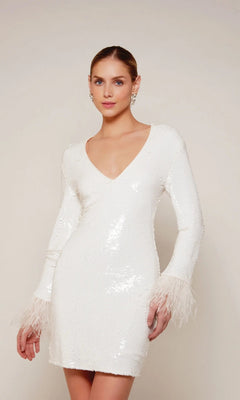 A woman stands against a plain light background, wearing the Alyce 70027 knee length white party dress featuring sequins and striking feather trim cuffs.