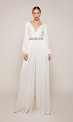 A woman models the Alyce 70031, a white satin-chiffon formal jumpsuit with long sleeves, wide legs, a deep V-neckline, and jeweled belt, set against a plain light background.