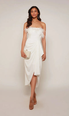 A woman in an ivory Alyce 70034 knee-length semi-formal dress with feather accents holds a small clutch and stands in sandals against a plain background.