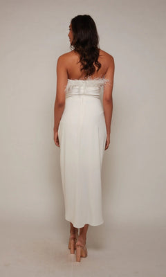 Woman facing away from the camera in a white, strapless Alyce 70034 knee-length semi-formal dress with feather accents, set against a plain background.