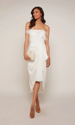 A woman poses against a plain light background wearing the Alyce 70034 knee length semi-formal strapless white dress with feather accents, complemented by a beige clutch and nude heels.
