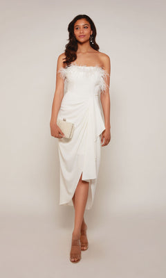 A woman poses against a plain background in the Alyce 70033 knee-length, white off-the-shoulder semi-formal dress with feather accents, styled with a small clutch and nude heels.