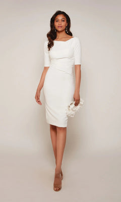 A woman wears the Alyce 70036 knee-length semi-formal dress in ivory, featuring an asymmetrical high neckline and half sleeves, and holds a white clutch against a plain light background.
