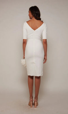 A woman faces away in the Alyce 70036 Knee Length Semi-Formal Dress, featuring a deep V-back and short sleeves. She holds a small white accessory and wears nude heels—ideal for cocktail parties, set against a plain background.