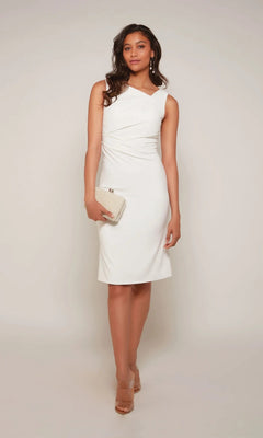 A woman stands against a plain light background wearing the Alyce 70037 knee-length white party dress, nude heels, and holding a textured beige clutch.