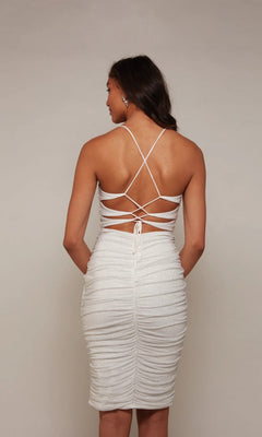 A woman models the Alyce 70039 knee-length semi-formal dress—form-fitting, white, with ruching and an open back featuring crisscross spaghetti straps—standing against a plain light backdrop.