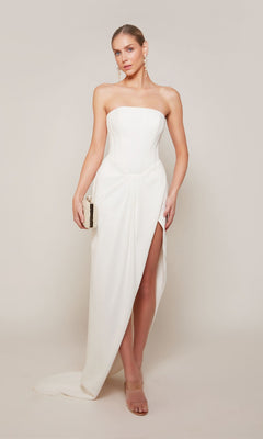 A woman wearing the Alyce 70042 long formal dress with a strapless corset bodice and thigh-high slit sheath skirt, holding a small clutch against a plain light background.