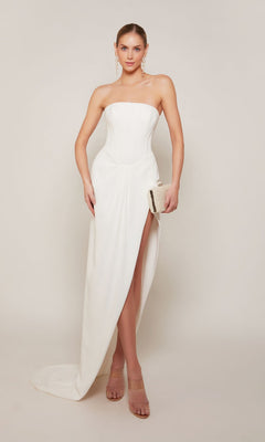 A woman in the Alyce 70042 long formal dress—strapless, white, with a corset bodice and thigh-high slit—stands against a plain background holding a clutch, wearing nude heels and drop earrings.