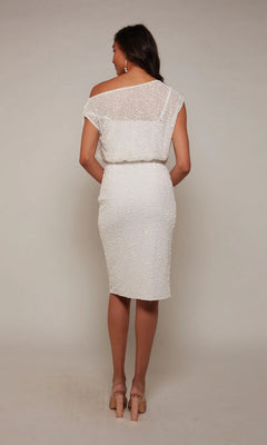 A woman stands with her back to the camera, wearing the Alyce 70046 knee-length white party dress featuring a sequin texture, short sleeves, and nude high-heeled shoes for an elegant, modern look.