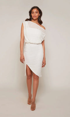 A woman stands against a plain background wearing the Alyce 70046 Knee Length White Party Dress, featuring an asymmetrical hem and one-shoulder neckline, styled with nude heels.