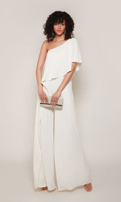 A woman stands against a light background wearing the Alyce 70049 long formal jumpsuit in ivory and holding a silver clutch bag.
