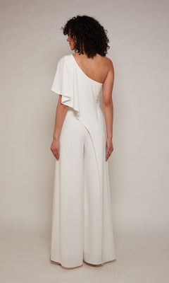 A person stands facing away, wearing the Alyce 70049 Long Formal Jumpsuit—a white, one-shoulder design with wide legs and an asymmetrical sleeve—creating an elegant cocktail look against a plain background.