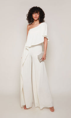 A woman wears the Alyce 70049 Long Formal Jumpsuit with an off-shoulder wide-leg design, holding a small silver clutch and standing in front of a plain light background.