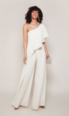 A woman poses against a plain background wearing the Alyce 70049 Long Formal Jumpsuit, a chic white one-shoulder outfit, and holds a small clutch bag.